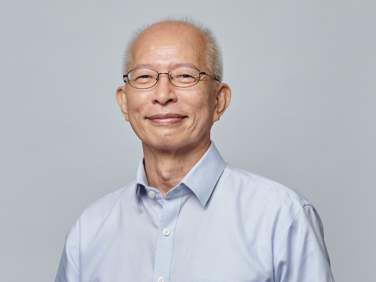 Dr Vincent Chua Cheng Huat in a light blue shirt smiling with one hand on his abdomen and the other in his pocket against a plain grey background